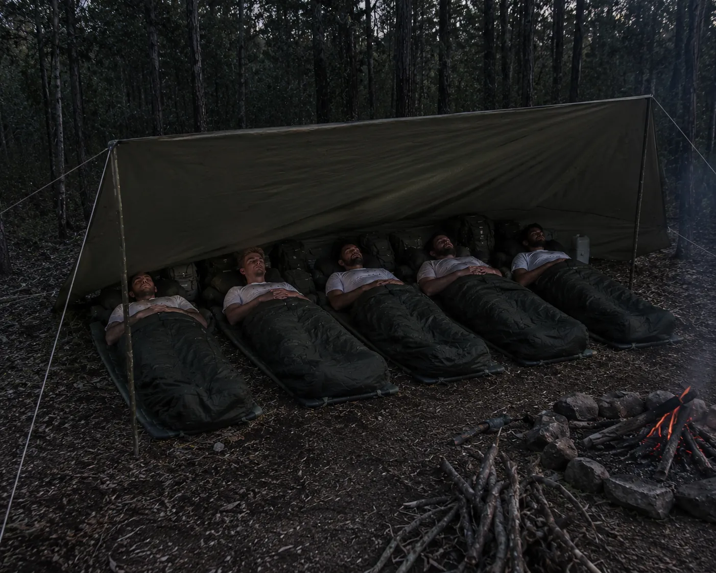 Participants sleeping in the field under a tarp shelter beside a campfire